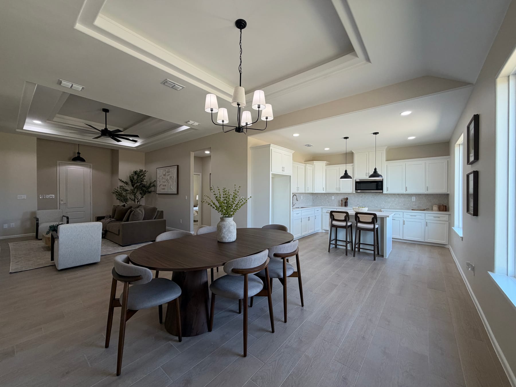 Dining table under a chandelier, with the living room to the left, the kitchen and island beyond, and framed art and windows along the right wall
