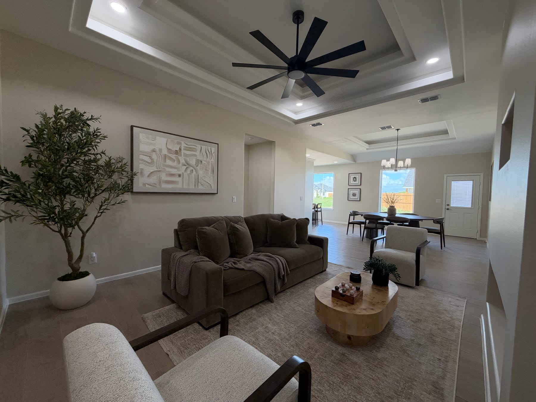 Living room under a tray ceiling with a black ceiling fan, staged with a sofa and armchairs, open to the dining area and a back door
