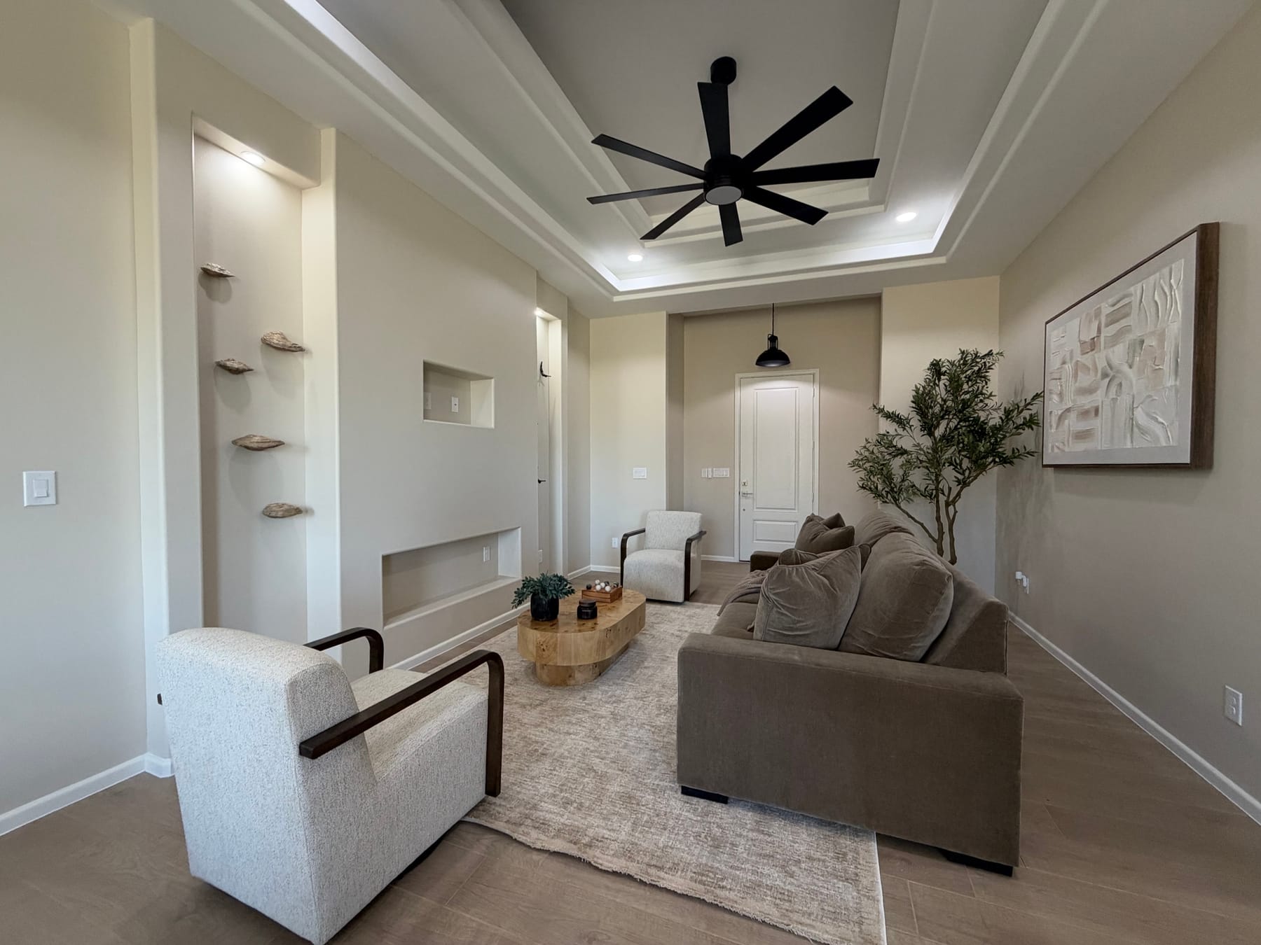 Living room with recessed wall niches and floating shelves under a tray ceiling with a fan, looking toward a door at the far end