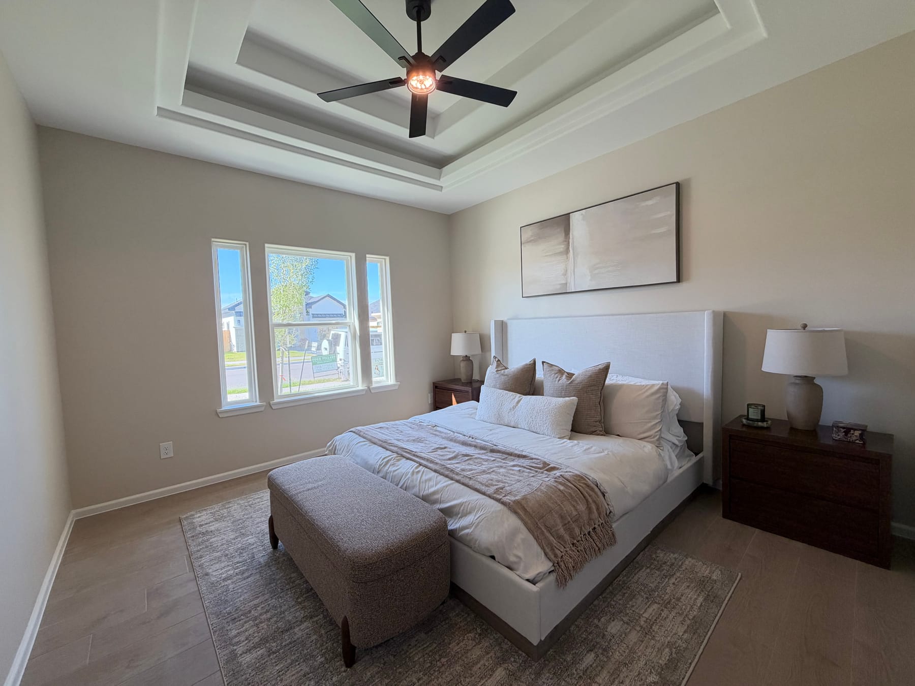 Primary bedroom under a tray ceiling with a fan, staged with a bed and an upholstered bench beneath a pair of windows