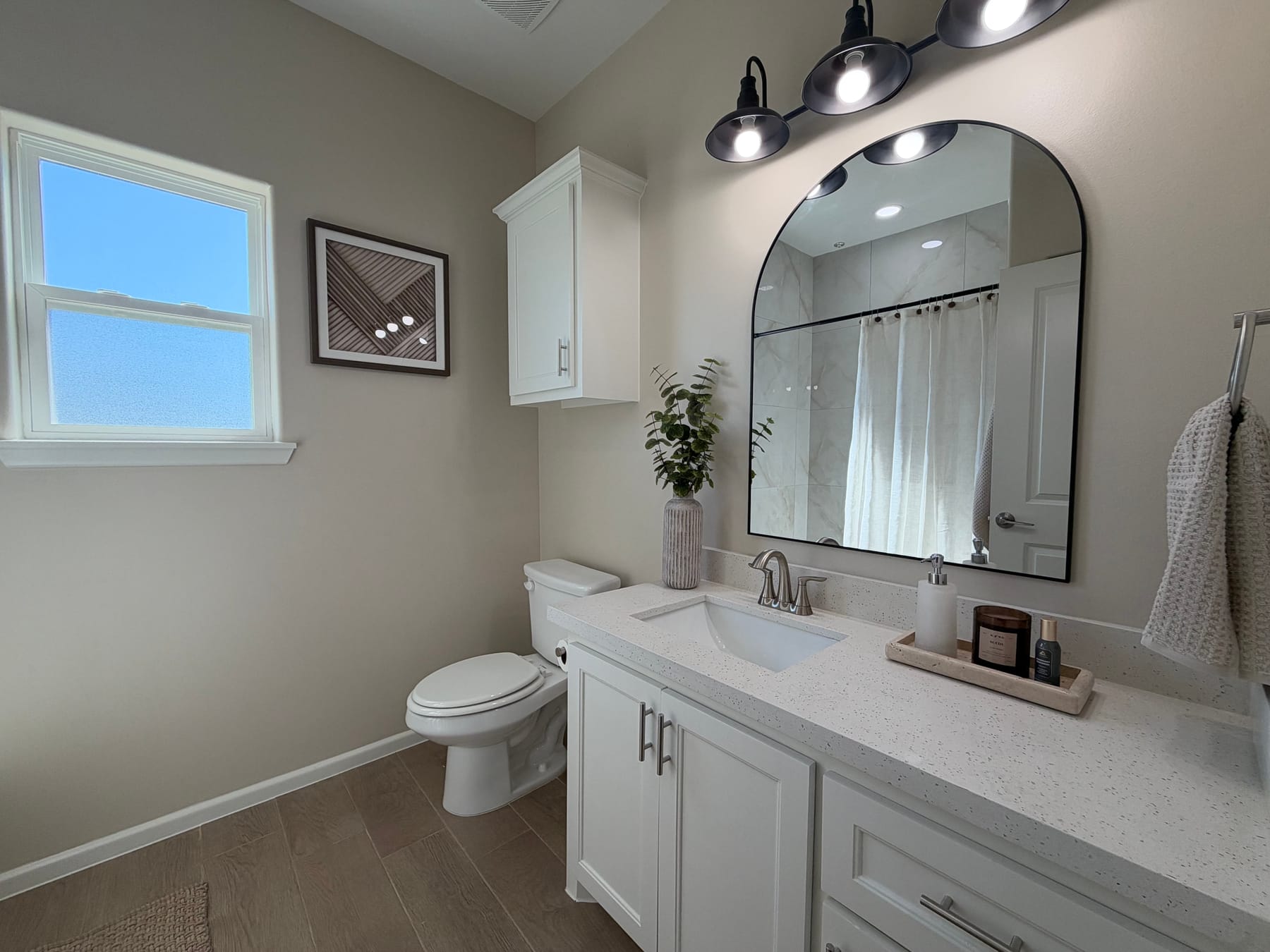 Hall bathroom with a single vanity, an arched black-framed mirror, a window and a cabinet above the toilet