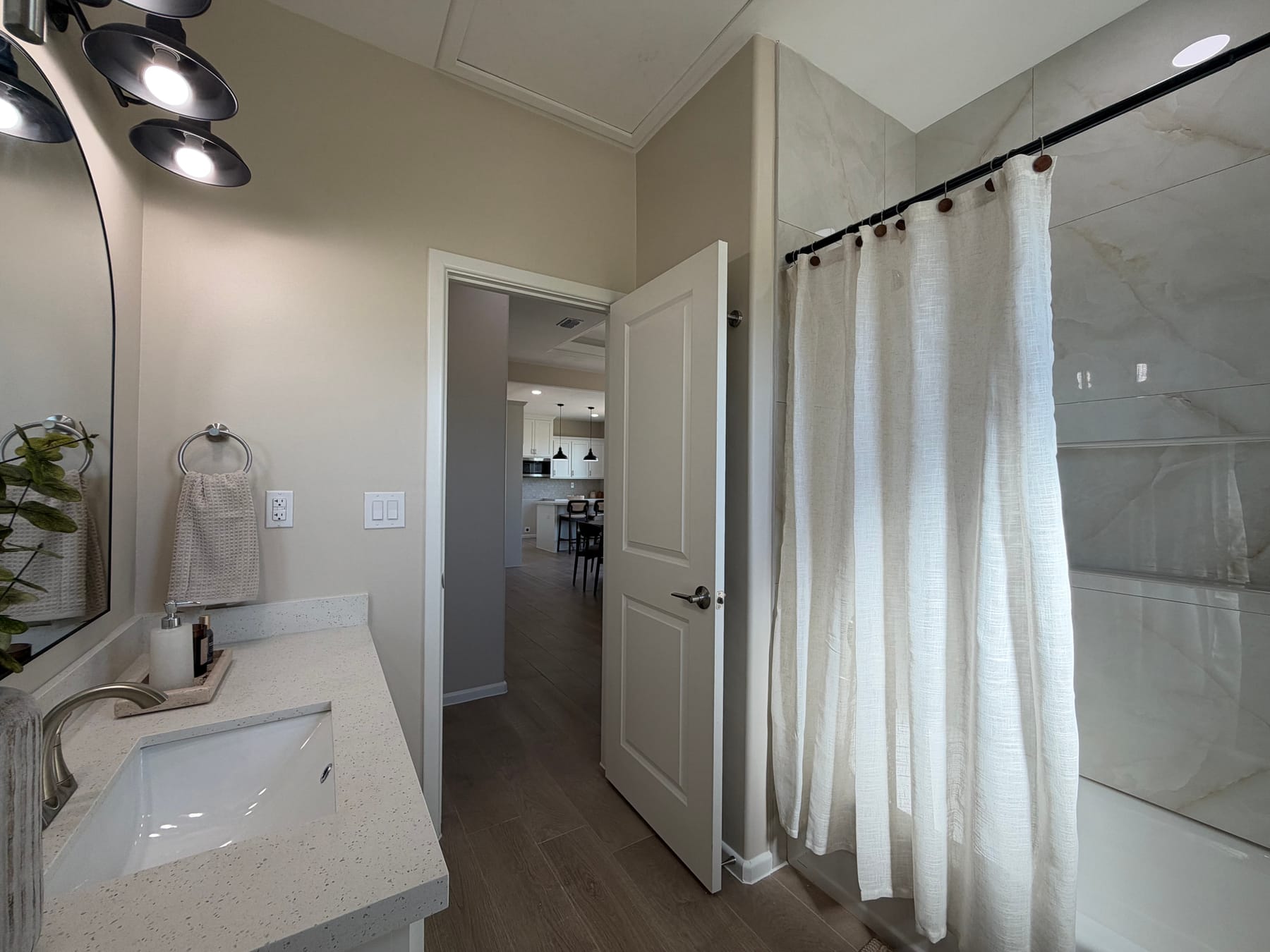 The hall bathroom's vanity beside a curtained tub and shower with marble-look tile, its door open to the hallway and kitchen